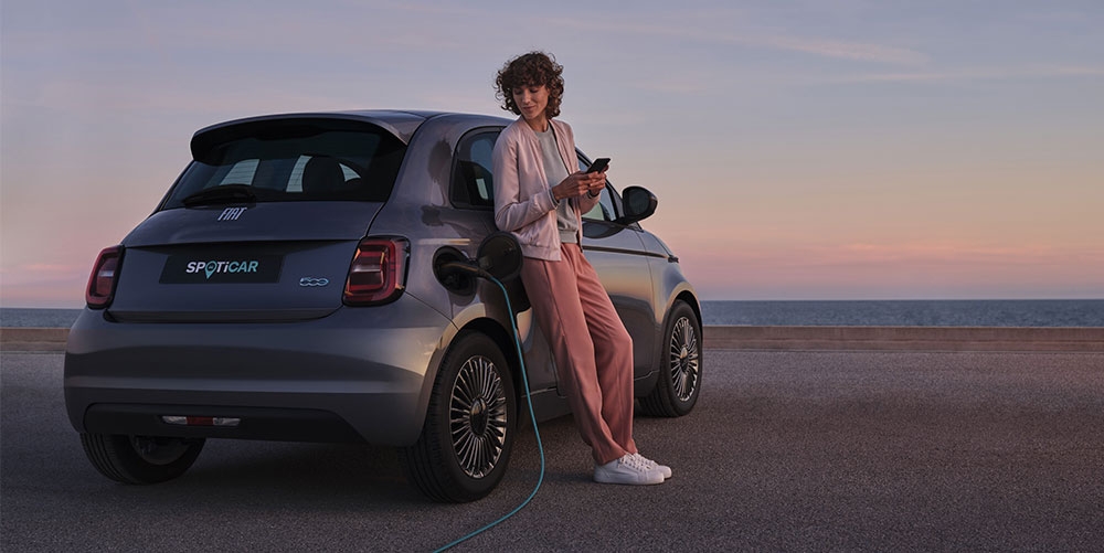 Lady leaning on FIAT charging her new SPOTICAR car